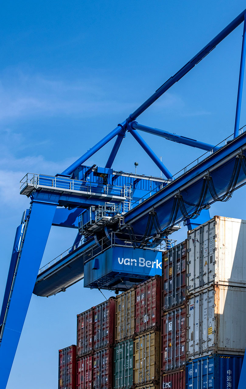 A large blue industrial crane is seen lifting and organizing colorful shipping containers at a shipping yard. The sky is clear and blue, providing a contrasting backdrop to the machinery and containers. The containers are stacked high, indicating an active and busy shipping operation.