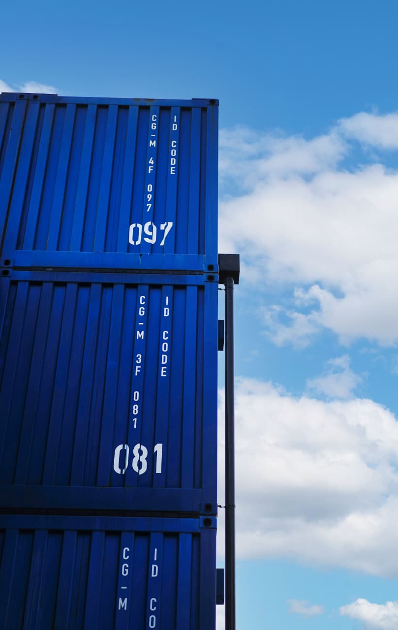 A close-up view of two blue shipping containers stacked on top of each other against a bright sky with white clouds. The containers are marked with white identification codes, "097" on the upper container and "081" on the lower one, emphasizing their organized and systematic nature. The image captures the scale and efficiency of containerized cargo, set against a backdrop of a clear, open sky, symbolizing the vastness and potential of global trade.