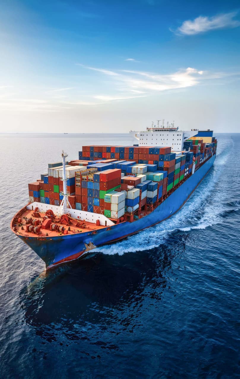 A large cargo ship, fully loaded with multicolored shipping containers, sails through calm, deep blue waters under a clear sky. The ship's bow is cutting through the water, creating white waves on either side. The vessel's deck is stacked high with containers, indicating a robust load. The image captures the scale and efficiency of maritime shipping, set against a backdrop of serene ocean and sky.